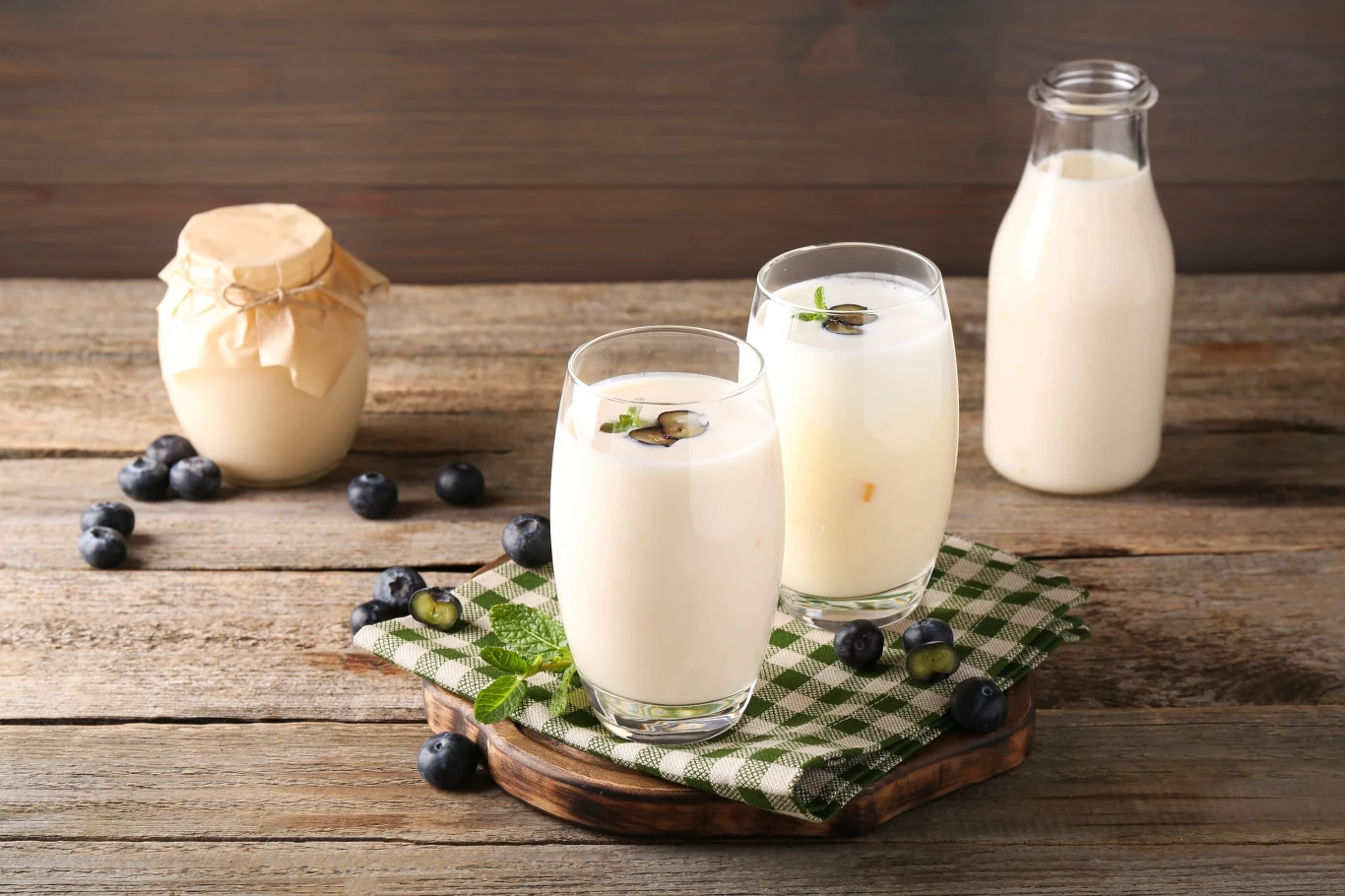 Maślanka czy kefir? Ekspert odpowiada, co lepiej działa na jelita Szklanki z kefirem i maślanką stojące na drewnianym stole obok świeżych owoców i produktów mlecznych.