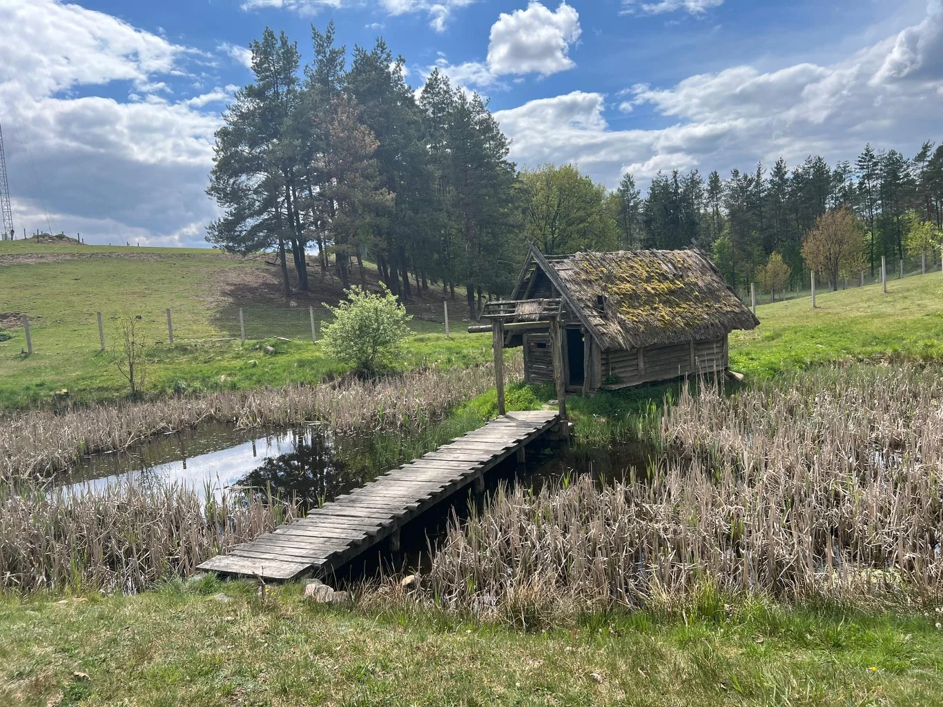 Stara drewniana chatka z mchem na dachu stoi tuż przy stawie, do którego prowadzi wąski pomost, otoczony suchymi trzcinami i trawą, w tle zalesione wzgórze pod pochmurnym niebem.