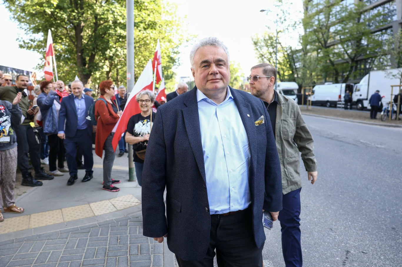 Tomasz Sakiewicz wezwany na przesłuchanie do prokuratury Tomasz Sakiewicz w garniturze idzie chodnikiem z grupą osób, część z biało-czerwonymi flagami, w tle drzewa