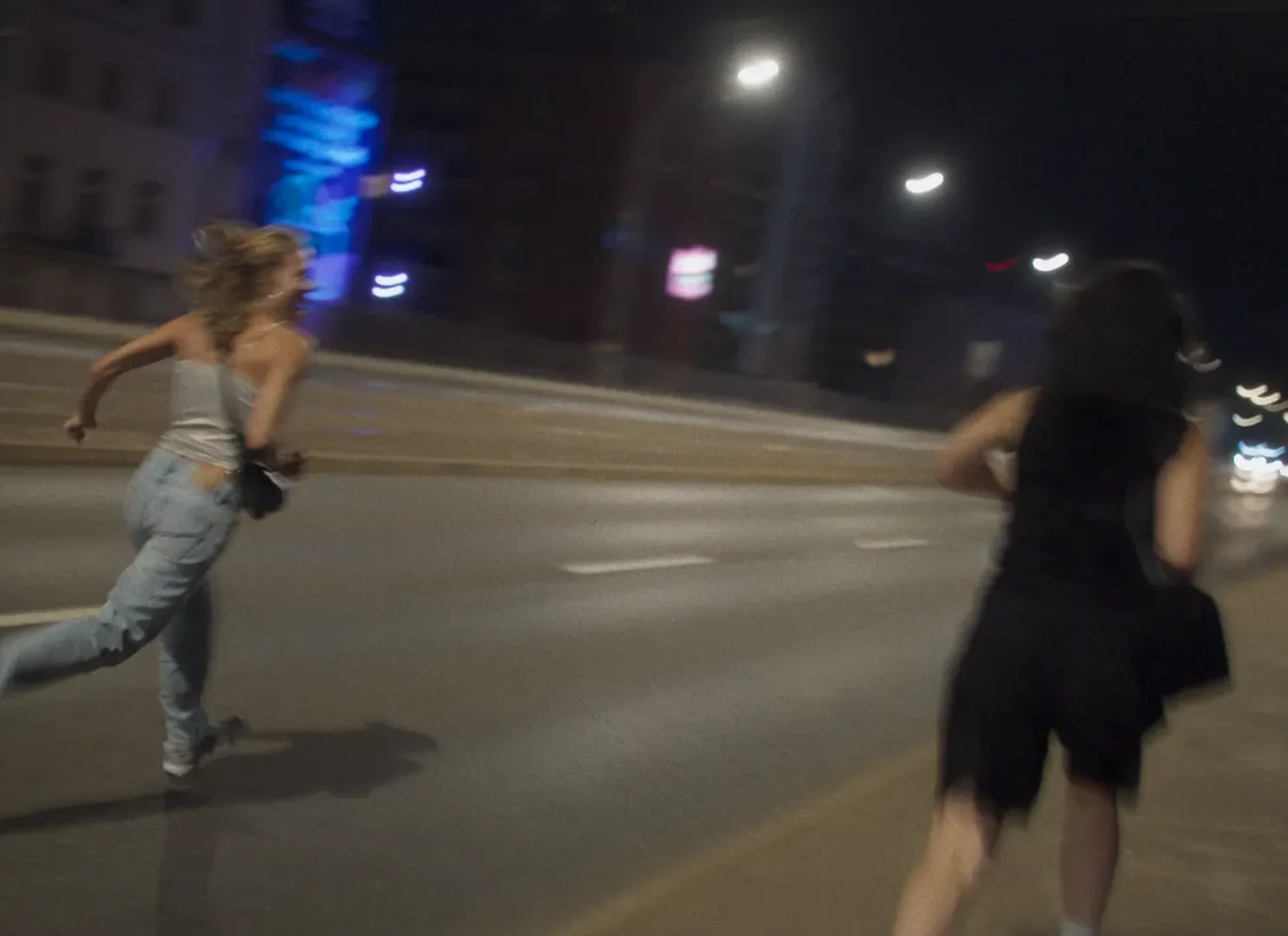 Dwie osoby biegną po pustej, oświetlonej ulicy nocą, jedna w dżinsach i szarej koszulce, druga w ciemnej sukience.