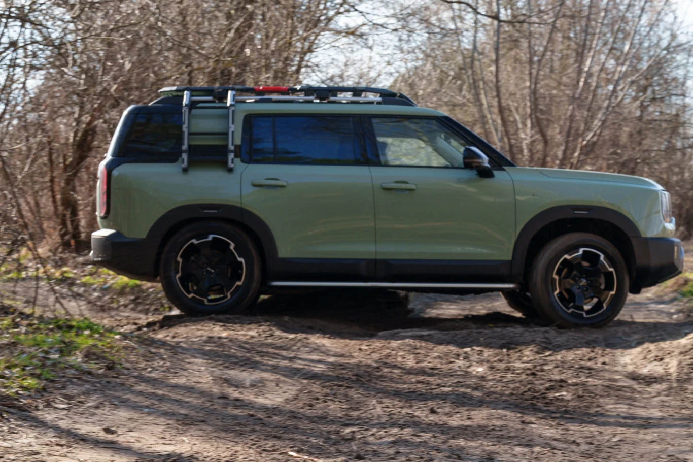 Jasnozielony SUV z relingami dachowymi i czarnymi felgami pokonuje terenową drogę otoczoną bezlistnymi drzewami.