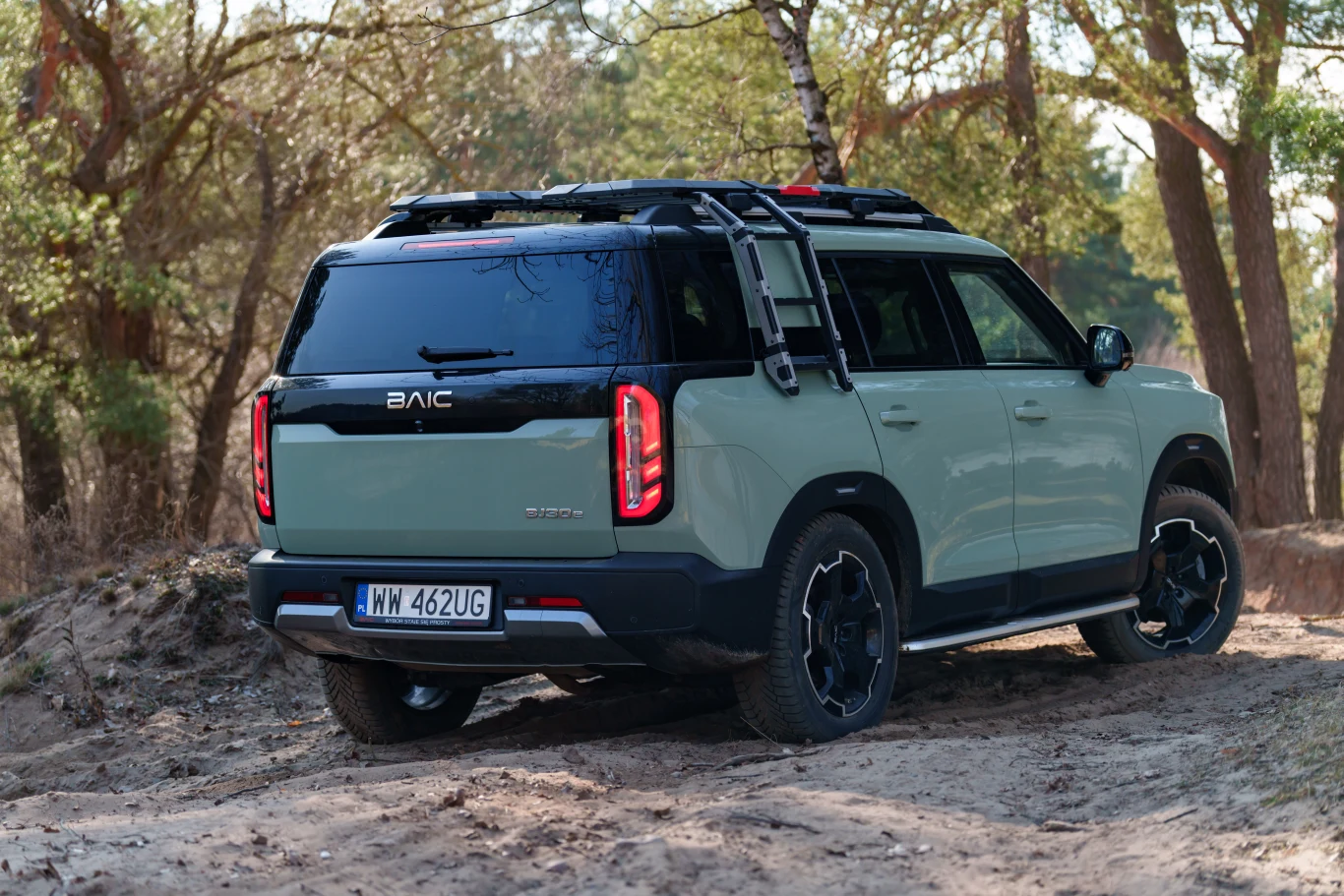 SUV marki BAIC o jasnej karoserii i czarnym dachu, wyposażony w bagażnik dachowy oraz dodatkowe relingi, stoi na piaszczystej, leśnej drodze otoczonej drzewami.