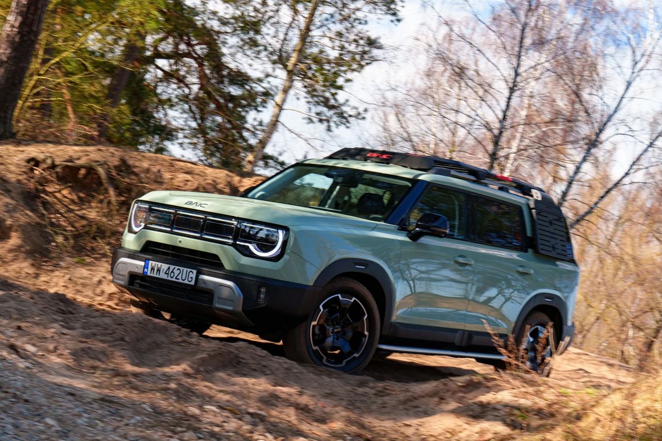 Jasnozielony samochód terenowy z czarnymi dodatkami pokonuje piaszczysty, leśny teren, podkreślając swoje możliwości off-roadowe.