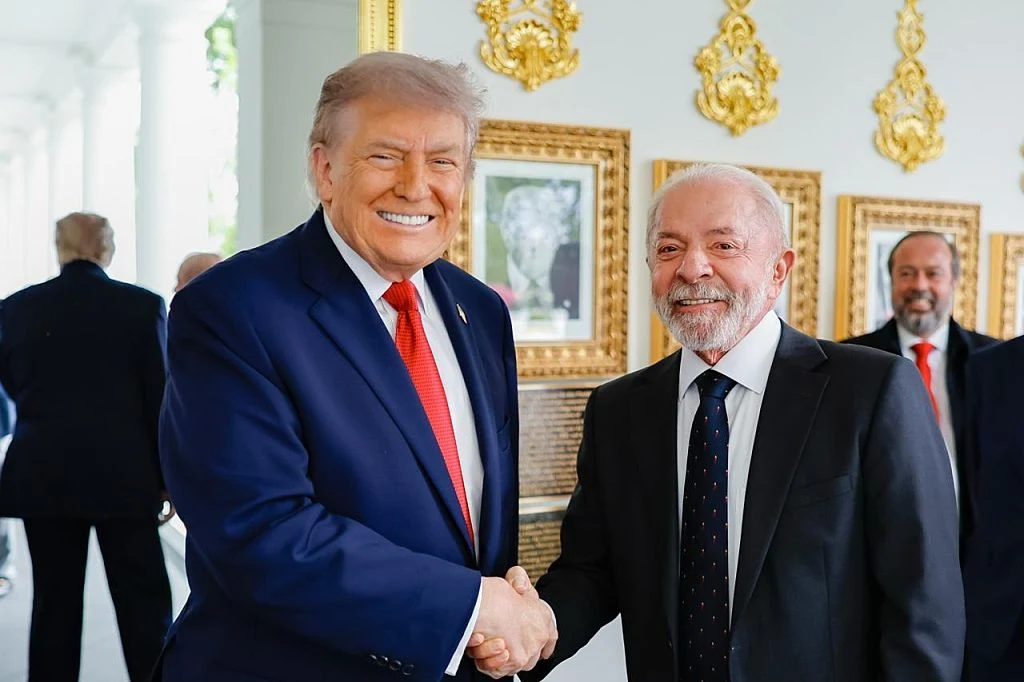 Dwóch elegancko ubranych mężczyzn w garniturach ściska sobie dłonie i uśmiecha się, na tle złotych ram i ozdobnych dekoracji na ścianie.