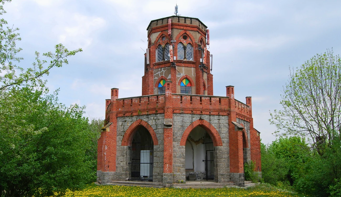 Ceglany budynek z ośmiokątną wieżą, gotyckimi łukami i barwnymi oknami, otoczony bujną zielenią oraz żółtymi kwiatami na trawniku.