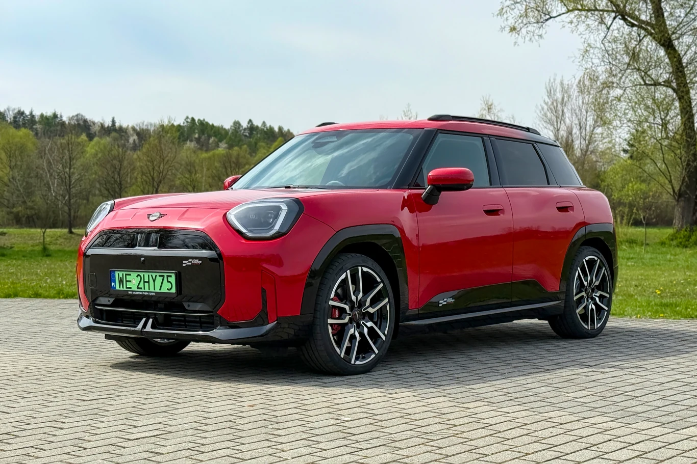 Czerwony SUV MINI stojący na brukowanym parkingu, wyposażony w duże aluminiowe felgi i przyciemniane szyby, z widoczną zieloną tablicą rejestracyjną i czarnymi akcentami karoserii.