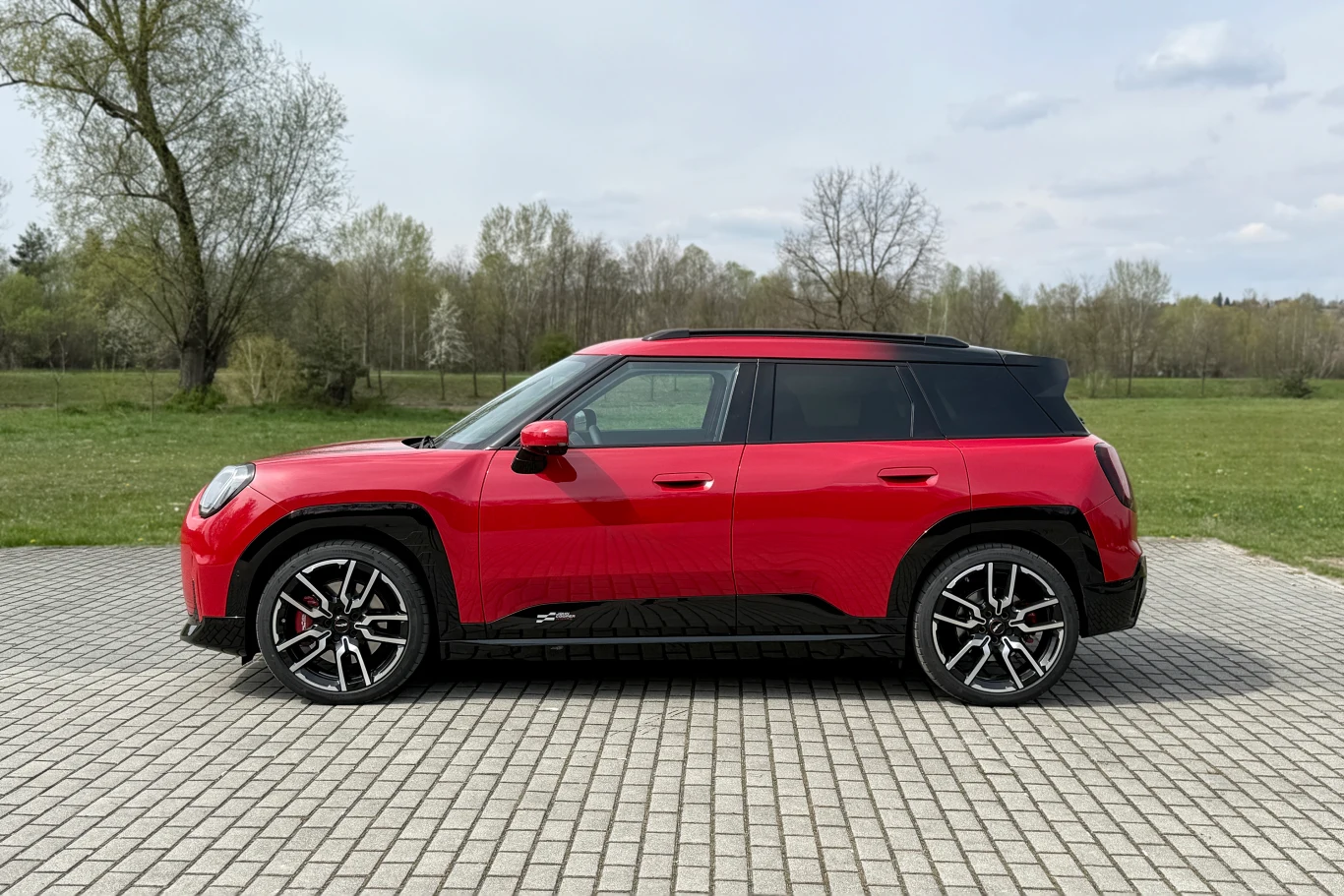 Czerwony samochód typu SUV zaparkowany na kostce brukowej, w tle wiosenna zieleń i rzadko rozstawione drzewa.