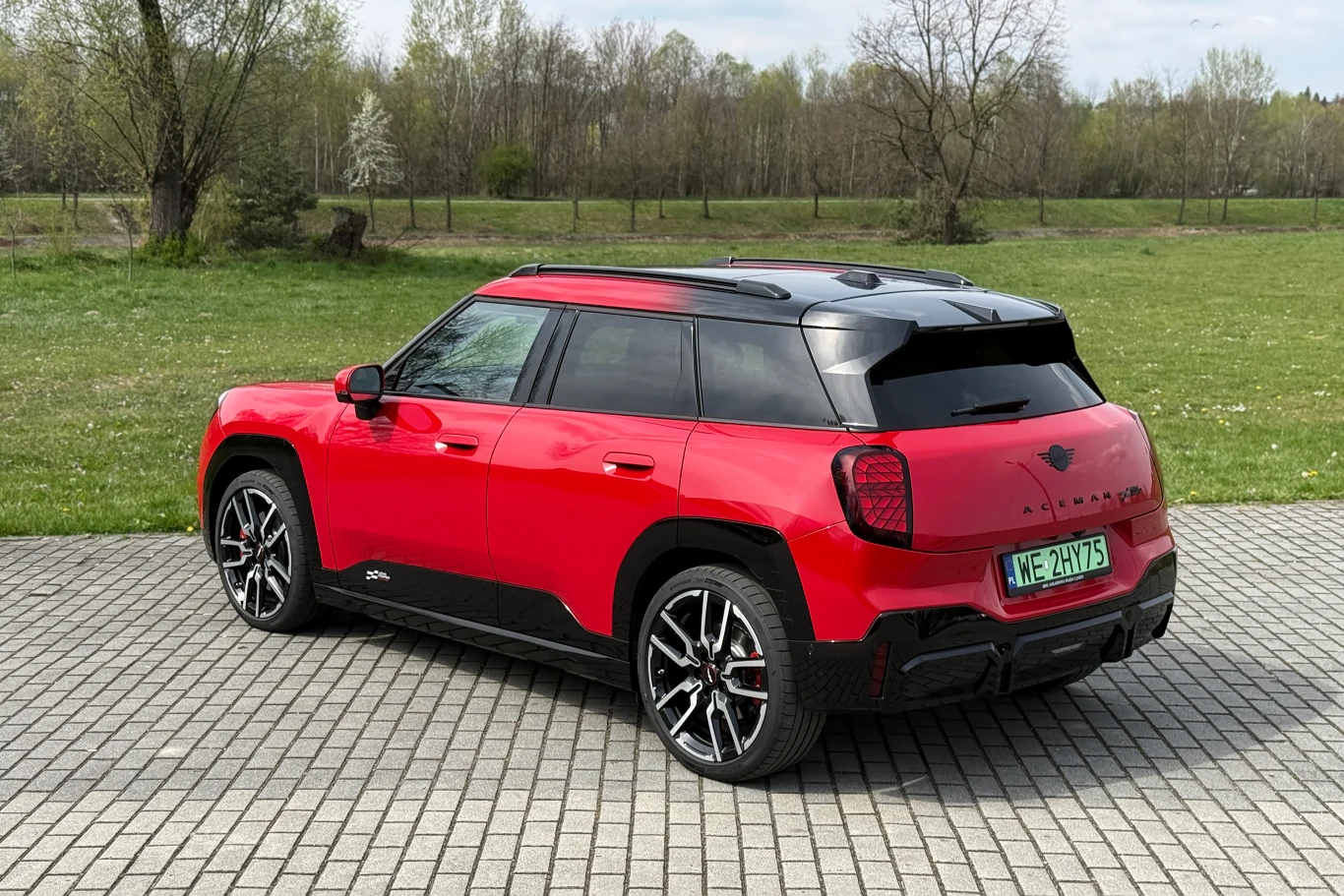 Czerwony samochód MINI Countryman o nowoczesnym designie stoi na kostce brukowej, otoczony zielonym trawnikiem i drzewami wiosną.