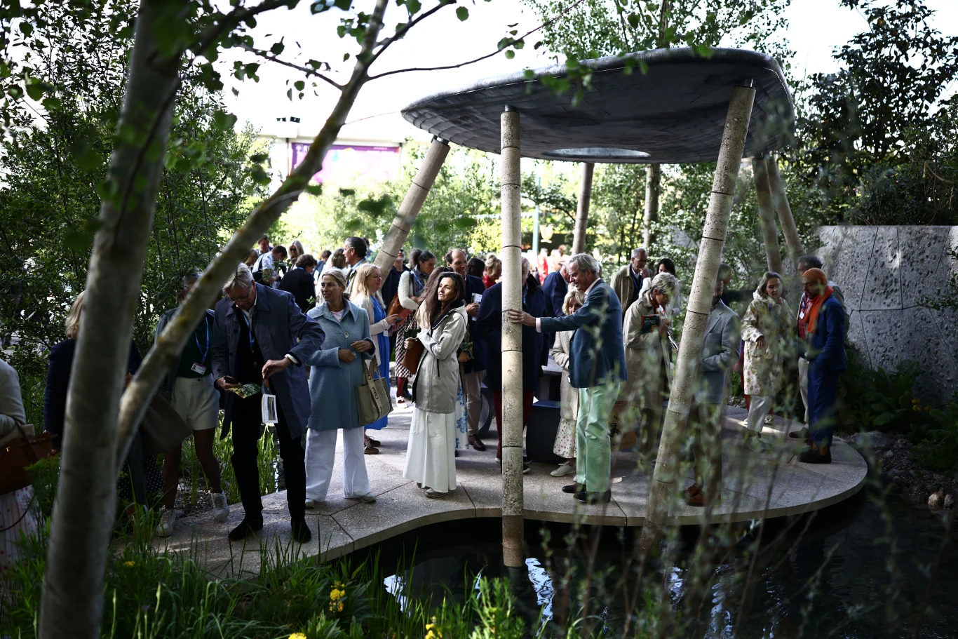 Wiele z ogrodów na Chelsea Flower Show stawia na ekologię i ponowne wykorzystanie zasobów Grupa elegancko ubranych osób zgromadzona pod dużą, okrągłą altaną w ogrodzie otoczonym zielenią i niewielkim zbiornikiem wodnym.