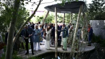 Grupa elegancko ubranych osób zgromadzona pod dużą, okrągłą altaną w ogrodzie otoczonym zielenią i niewielkim zbiornikiem wodnym.