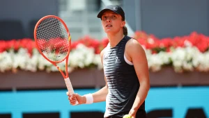 Tenisistka ubrana w sportową odzież, z rakietą w prawej ręce i piłką tenisową w lewej, stoi na korcie podczas meczu, w tle widoczne kwiaty.