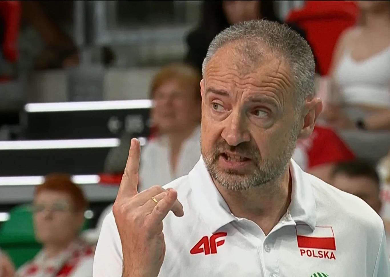 Nikola Grbić Mężczyzna w białej koszulce z napisem „Polska” podnosi palec wskazujący, mając poważny wyraz twarzy, w tle widać rozmytych kibiców na trybunach.