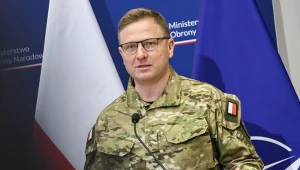 Mężczyzna w wojskowym mundurze z polską flagą na ramieniu przemawia przy mównicy, w tle widoczne flagi Polski i NATO oraz fragmenty logotypów Ministerstwa Obrony Narodowej.