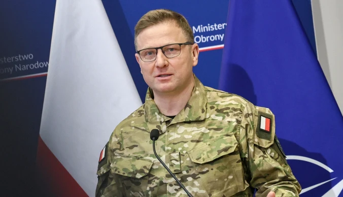 Mężczyzna w wojskowym mundurze z polską flagą na ramieniu przemawia przy mównicy, w tle widoczne flagi Polski i NATO oraz fragmenty logotypów Ministerstwa Obrony Narodowej.