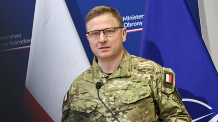 Mężczyzna w wojskowym mundurze z polską flagą na ramieniu przemawia przy mównicy, w tle widoczne flagi Polski i NATO oraz fragmenty logotypów Ministerstwa Obrony Narodowej.