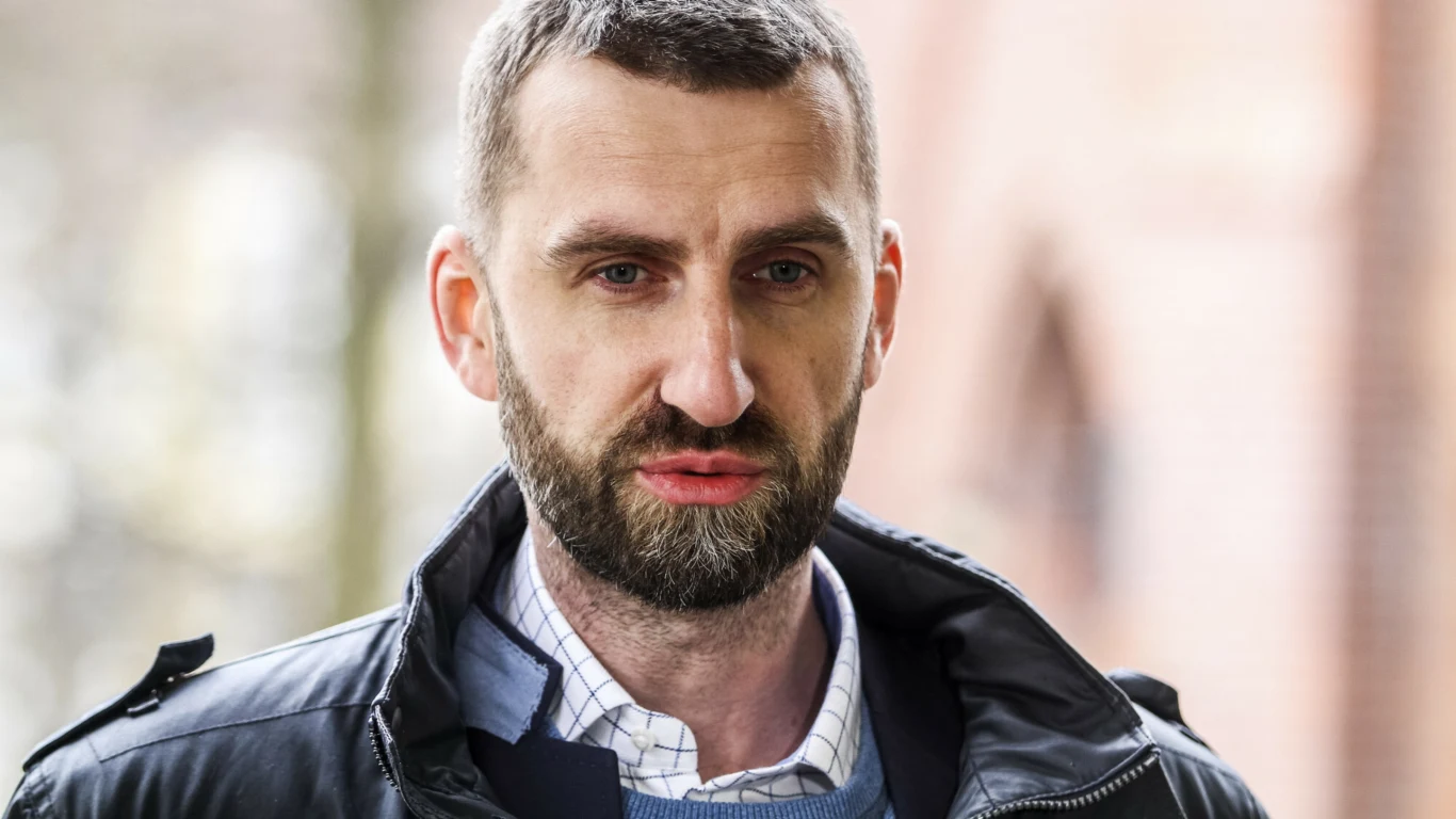 Mężczyzna z krótkimi włosami i brodą w ciemnej kurtce oraz niebieskim swetrze, na tle rozmazanej architektury i jasnych drzew.