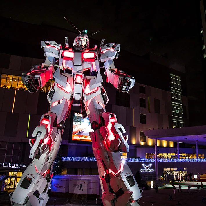 Wielka, podświetlana figura robota mecha stojąca przed nowoczesnym budynkiem, otoczona miejskim oświetleniem i zebranymi wokół ludźmi.
