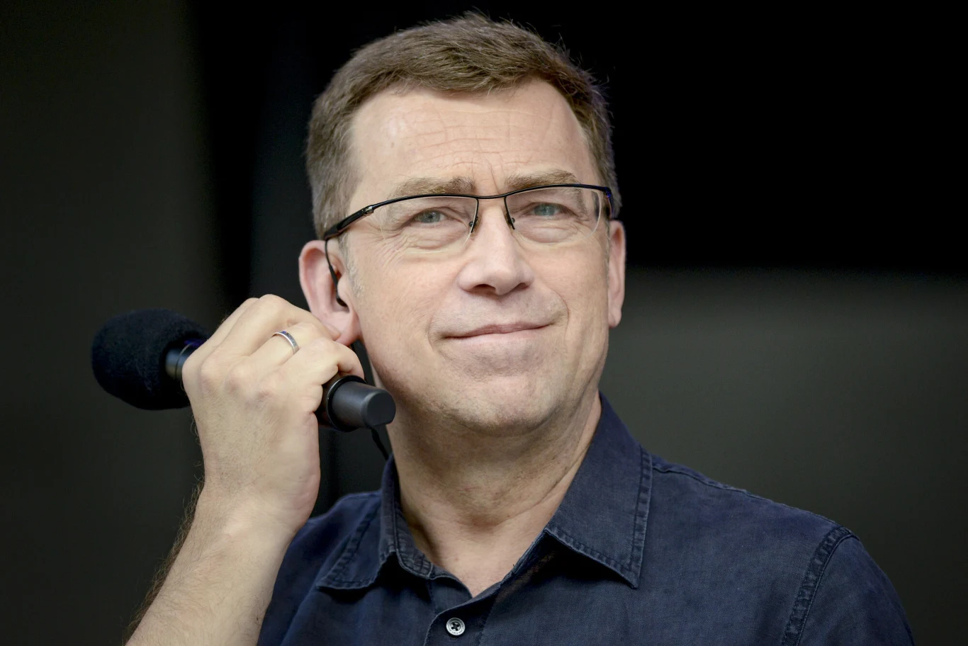 Maciej Orłoś Prezenter Maciej Orłoś w granatowej koszuli trzymający mikrofon.