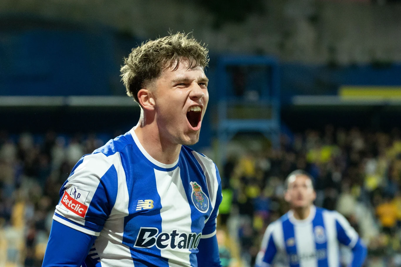 Oskar Pietuszewski Piłkarz w koszulce FC Porto wyraża silne emocje po akcji podczas meczu, w tle rozmyta sylwetka kolejnego zawodnika w takich samych barwach oraz widownia na stadionie.