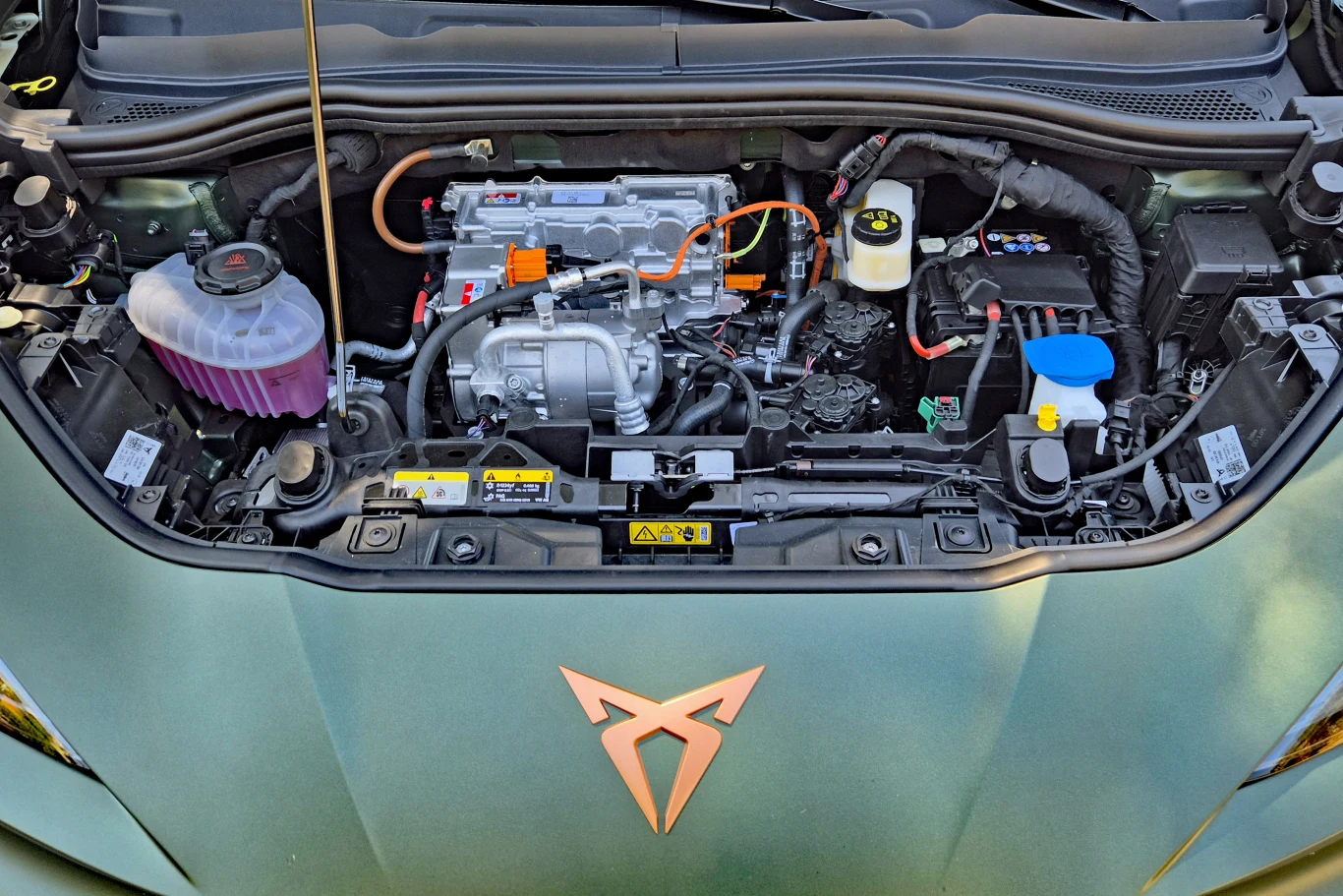 Komora silnika samochodu hybrydowego z widocznym silnikiem elektrycznym, akumulatorami oraz zbiornikami płynów eksploatacyjnych, na masce charakterystyczne logo marki Cupra.