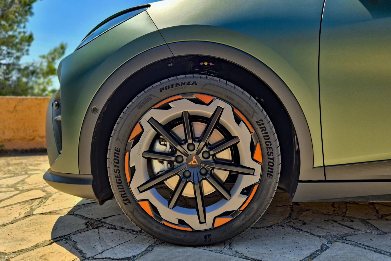 Nowoczesna felga aluminiowa z wyrazistymi, pomarańczowymi akcentami oraz opona Bridgestone Potenza zamontowana na przednim kole zielonego samochodu zaparkowanego na kamiennej nawierzchni.