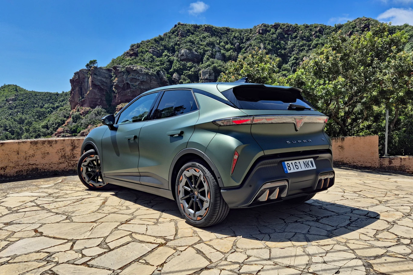 Nowoczesny, sportowy SUV w matowym zielonym kolorze z wyróżniającymi się tylnymi światłami LED, agresywnym dyfuzorem i czterema końcówkami układu wydechowego, zaparkowany na kamiennym tarasie w otoczeniu górskiej scenerii.