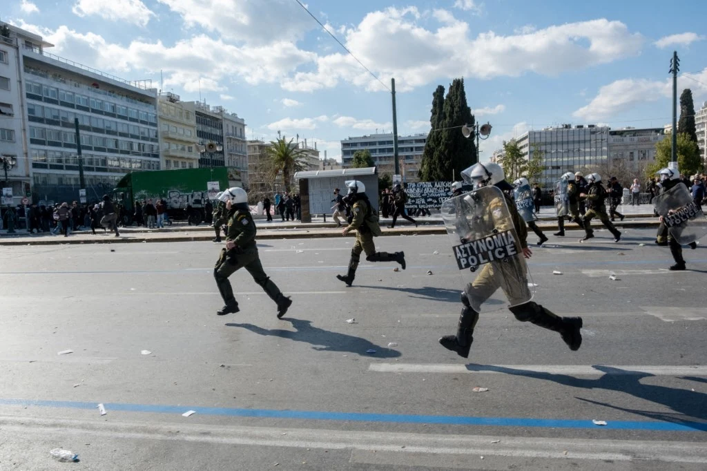 Grupa policjantów w pełnym rynsztunku ochronnym biegnących przez ulicę w kierunku zgromadzenia ludzi w centrum miasta; w tle nowoczesne budynki i drzewa.