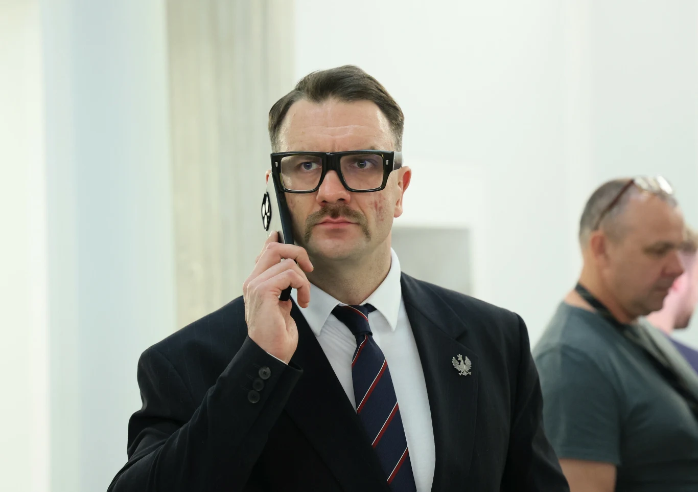 Mężczyzna w garniturze i okularach rozmawia przez telefon, w tle niewyraźna postać innego mężczyzny.