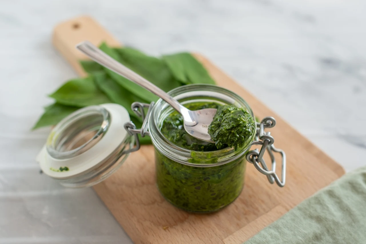 Słoik z zielonym pesto ustawiony na drewnianej desce, w środku łyżeczka z porcją sosu; w tle zielone liście.