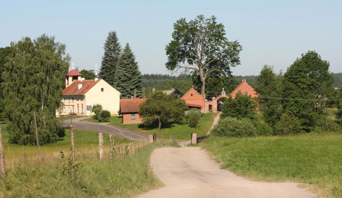 Kolekcja jasnych budynków gospodarczych z czerwonymi dachami otoczonych zielonymi drzewami i trawnikiem, na pierwszym planie polna droga prowadząca w kierunku zabudowań.