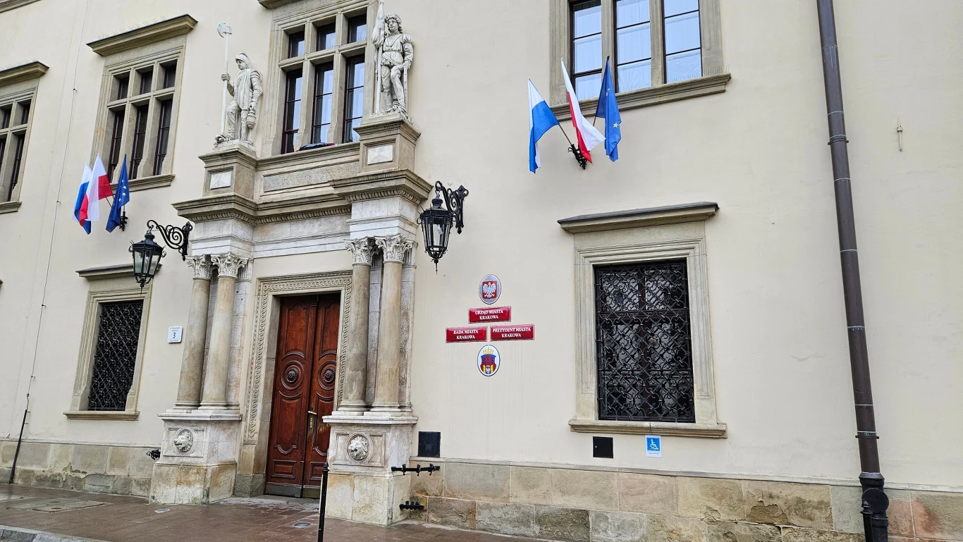 Urząd miasta Krakowa Wejście do historycznego budynku ozdobione rzeźbionym portalem, po bokach dwie kamienne figury, nad drzwiami umieszczone flagi oraz tablice z herbem i nazwą urzędu.