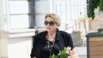 Kobieta w czarnym stroju, z okularami przeciwsłonecznymi i torebką na ramieniu, trzyma w dłoniach bukiet różowych kwiatów przewiązanych wstążką.