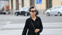 Kobieta w eleganckiej, czarnej sukience i okularach przeciwsłonecznych przechodzi przez ulicę miejską z torebką na łańcuszku przewieszoną przez ramię.