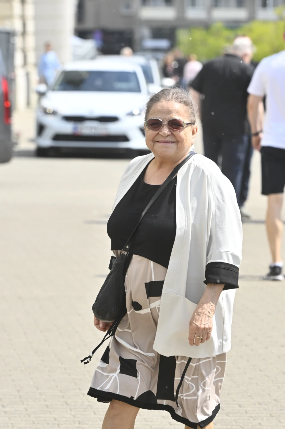 Grażyna Zielińska na pogrzebie Stanisławy Celińskiej Starsza kobieta w okularach przeciwsłonecznych ubrana w biało-czarną sukienkę i jasny żakiet spaceruje chodnikiem w słoneczny dzień.