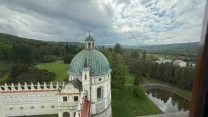 Barokowa kopuła kościoła otoczona zielenią, obok niewielki staw w kształcie podkowy, w tle rozległy krajobraz z lasami i polami.