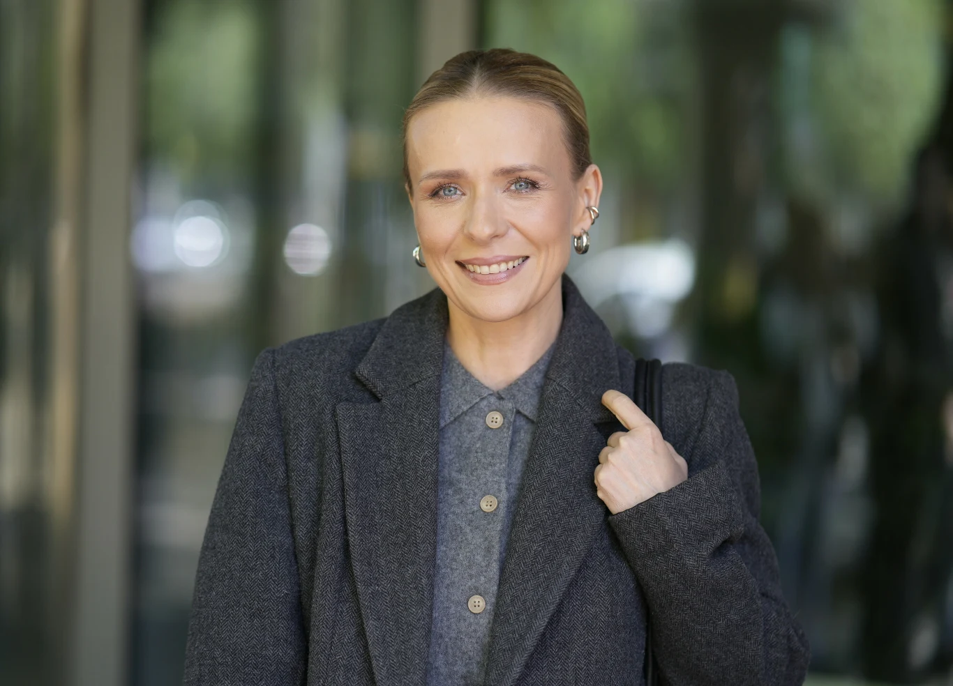 Zdaniem fanów, Aneta Zając wygląda dziś lepiej niż kiedykolwiek Uśmiechnięta kobieta w szarej, eleganckiej marynarce i kolczykach, trzymająca pasek swojej torby na ramieniu, stoi na tle nowoczesnego budynku.