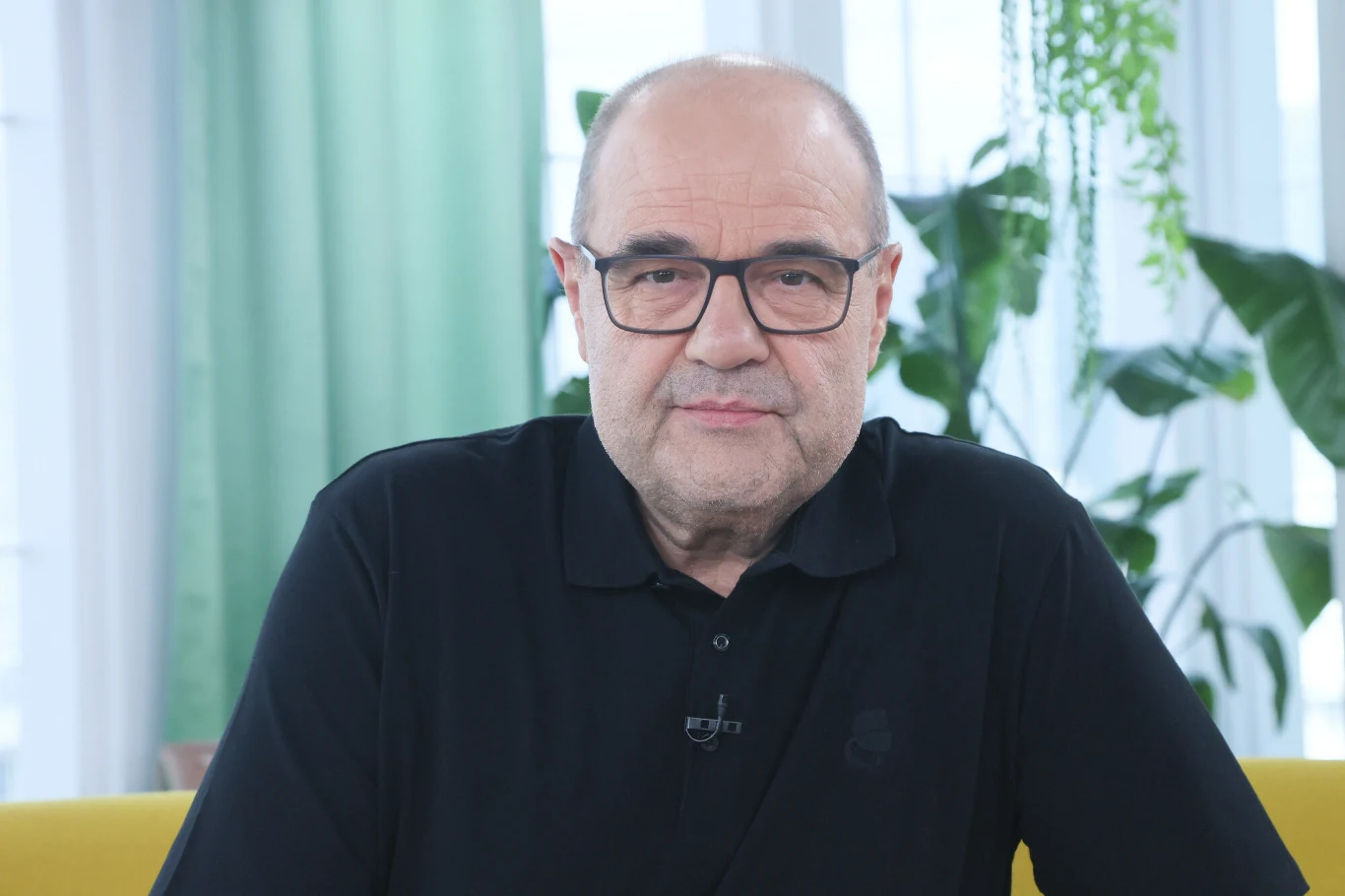 Cezary Żak zrzucił aż 30 kg i do dziś udaje mu się utrzymywać prawidłową masę ciała Starszy mężczyzna w okularach i czarnej koszulce z mikrofonem przypiętym do kołnierza, siedzący na żółtej sofie na tle dużych okien z widoczną zieloną roślinnością.