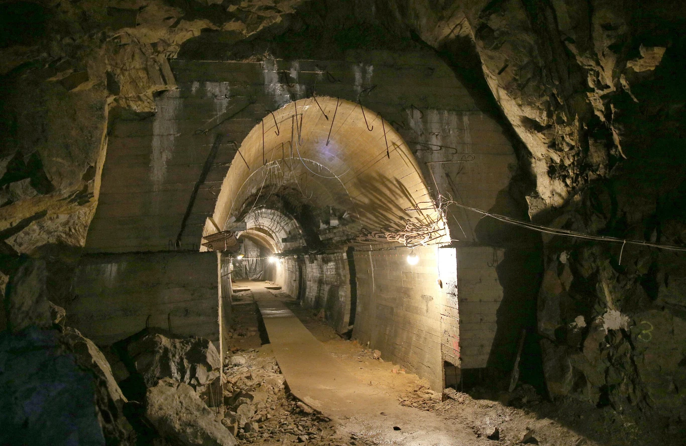 Betonowy korytarz w wykutym w skale tunelu, oświetlony lampami i poprowadzony w głąb góry, z widocznymi fragmentami instalacji elektrycznych na ścianach.