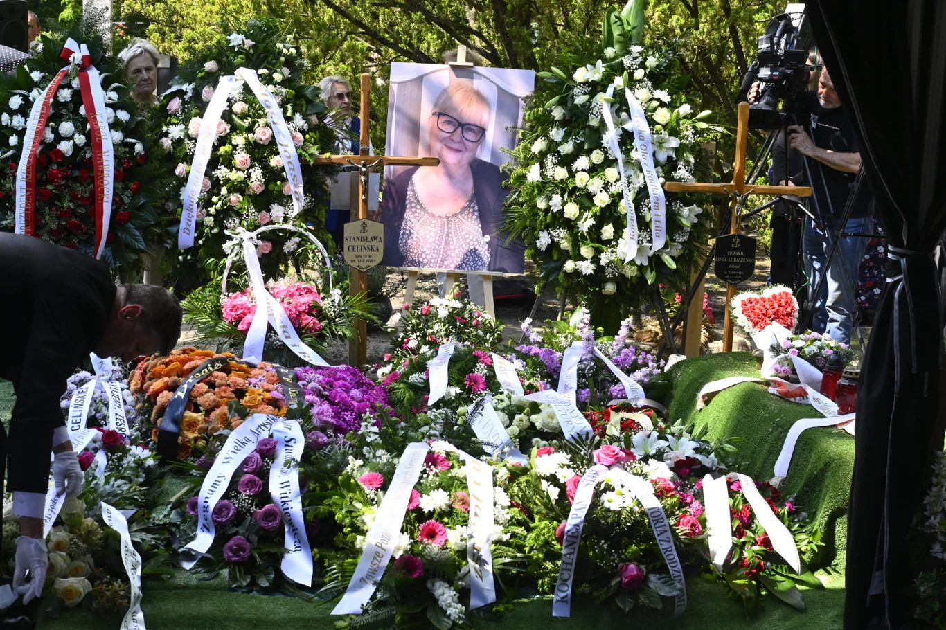 Portret zmarłej kobiety ustawiony na stojaku, duże wieńce i wiązanki kwiatów ozdobione biało-czarnymi wstążkami ułożone wokół grobu, zebrani żałobnicy stojący w tle.