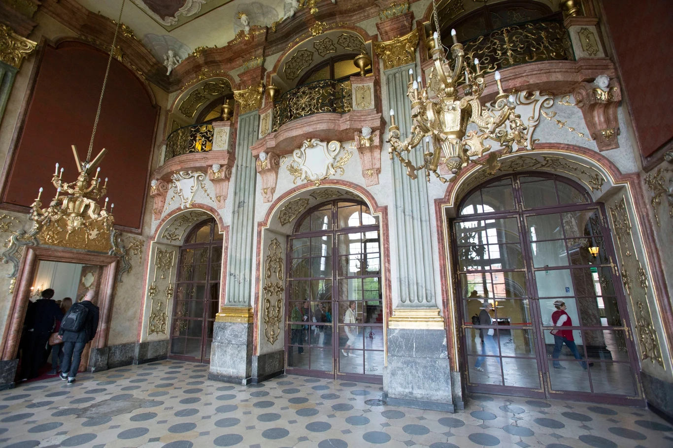 Bogato zdobione wnętrze z wysokimi oknami, złotymi ornamentami, żyrandolami oraz ozdobnymi balustradami, w którym przebywają osoby podziwiające architekturę.