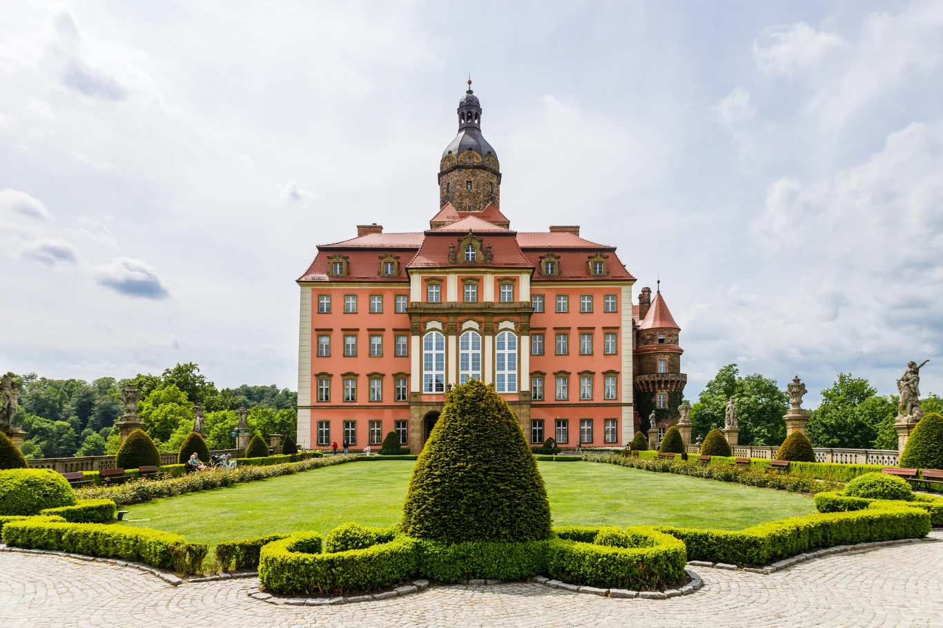 Różowy zamek o barokowej architekturze z wysoką wieżą centralną, otoczony starannie przystrzyżonymi krzewami i geometrycznym ogrodem na pierwszym planie.