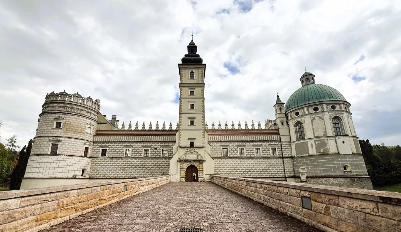 Renesansowy zamek z wysoką wieżą centralną, okrągłą basztą po lewej oraz charakterystyczną kopułą po prawej stronie, otoczony kamiennym murem i szeroką brukowaną drogą prowadzącą do wejścia.