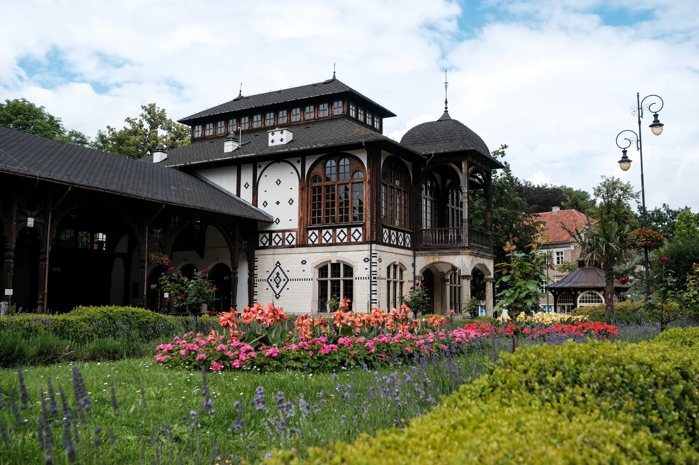 Zabytkowy budynek o wyraźnej drewnianej konstrukcji z dużymi oknami, otoczony kolorowymi rabatami kwiatowymi i zadbanym trawnikiem, w tle widoczne drzewa oraz elementy parkowej architektury.
