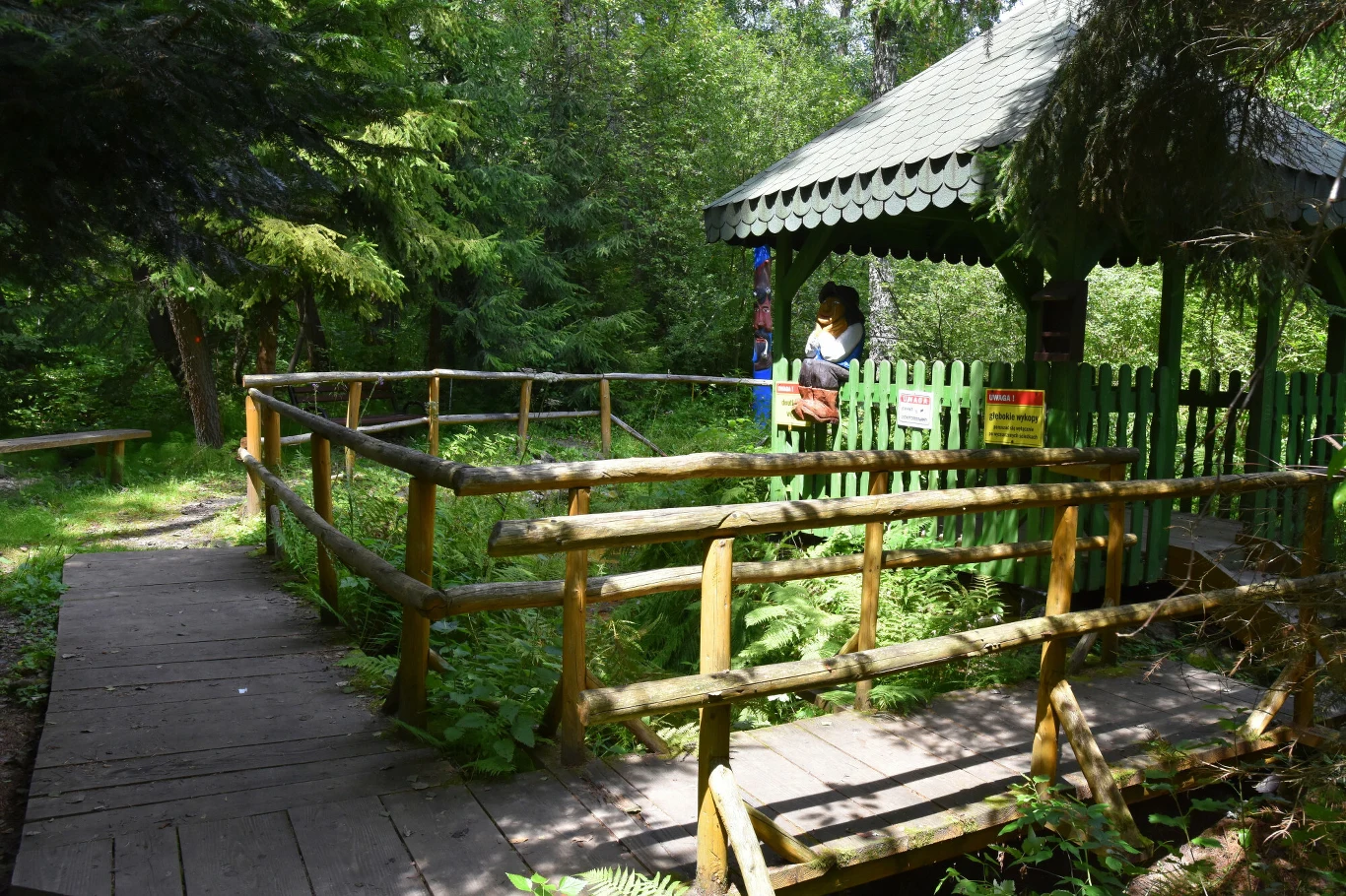 Drewniana altana otoczona zielonym płotkiem i balustradą pośród gęstego, leśnego krajobrazu, z figurą w stroju ludowym stojącą pod zadaszeniem.