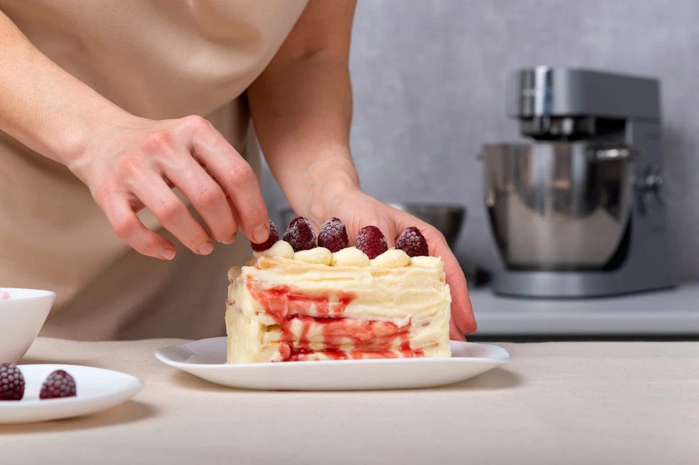 To tiramisu z malinami pokochają wszyscy goście Kawałek tiramisu z malinami na białej paterze, dekorowany świeżymi malinami przez osobę w jasnym fartuchu.