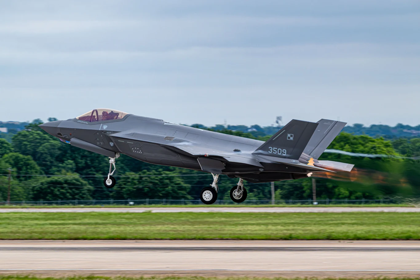 Wojskowy myśliwiec F-35 z polskimi oznaczeniami w trakcie startu, z widocznym podwoziem i silnikiem wytwarzającym płomień.