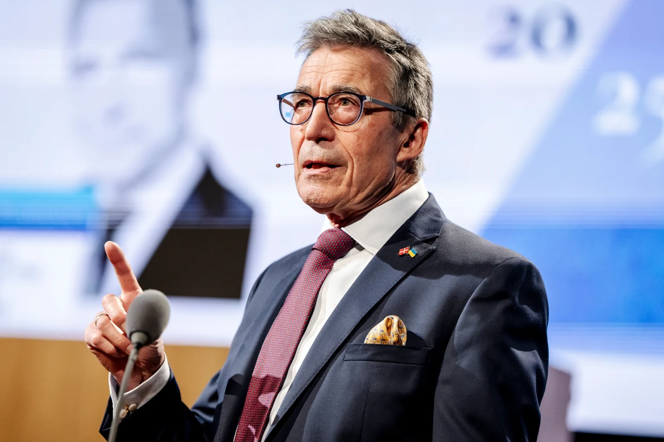 Były szef NATO i były premier Danii Anders Fogh Rasmussen Elegancko ubrany mężczyzna w garniturze i okularach, z przypiętym mikrofonem i drobną flagą w klapie, przemawia gestykulując przy mównicy na tle dużego ekranu z rozmazanym portretem.