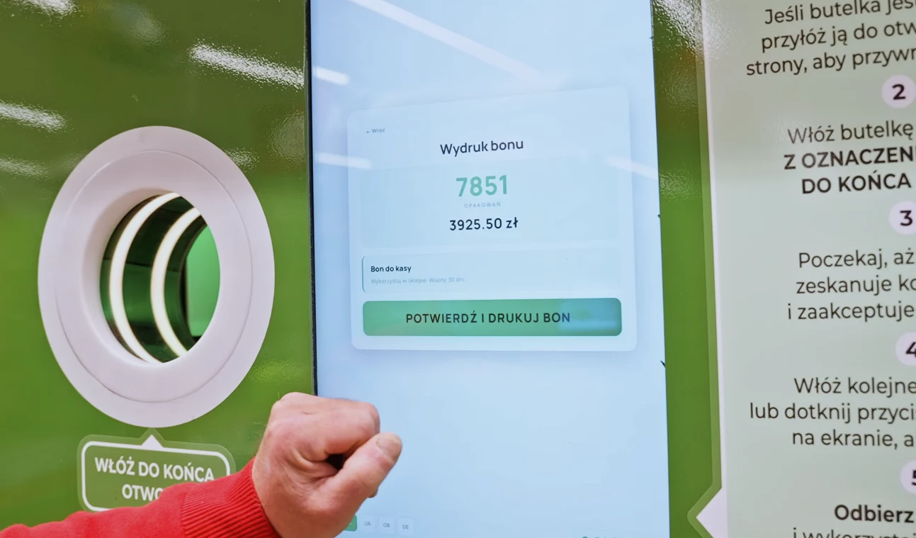 Interaktywny ekran automatu do przyjmowania butelek pokazujący wysoką kwotę bonu oraz licznik oddanych opakowań, obok widoczna ręka osoby potwierdzającej odbiór wydruku.