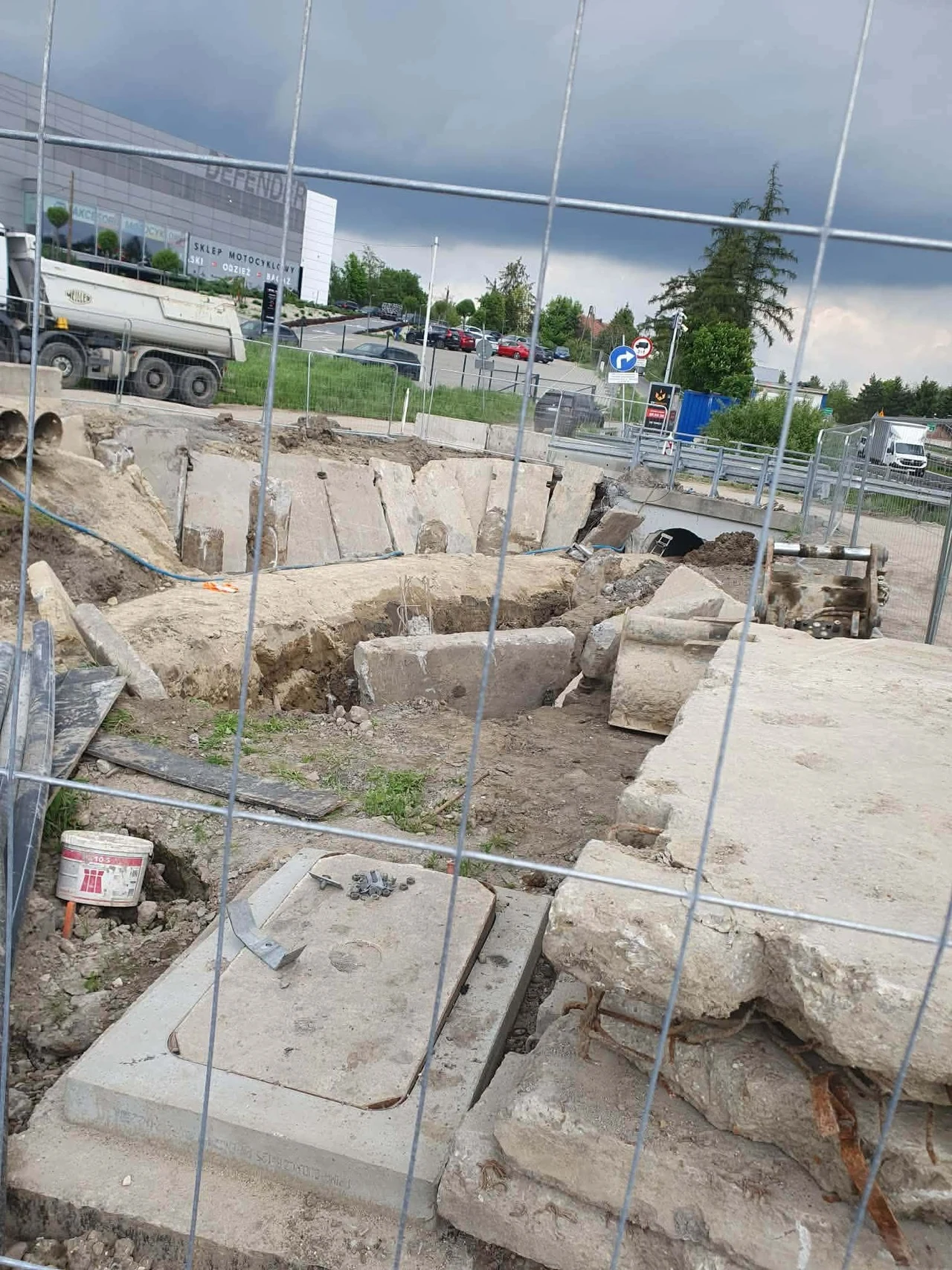 Rozkopany teren budowy z dużym wykopem oraz fragmentami betonowych płyt i rur, za ogrodzeniem budowlanym; w tle samochody i budynek biurowy.