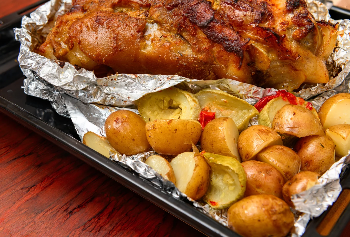 Pieczona golonka w złocistej, chrupiącej skórce podana z pieczonymi ziemniakami, wszystko ułożone na blasze wyłożonej folią aluminiową.
